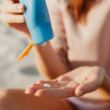 A person in a beige shirt applies sunscreen from a blue bottle onto their hand, with a blurred sandy beach background, evoking skin protection.