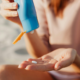 A person in a beige shirt applies sunscreen from a blue bottle onto their hand, with a blurred sandy beach background, evoking skin protection.