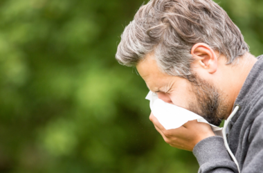 A man with gray hair is sneezing into a tissue outdoors. He is wearing a gray hoodie against a blurred green background, depicting allergy symptoms.