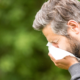 A man with gray hair is sneezing into a tissue outdoors. He is wearing a gray hoodie against a blurred green background, depicting allergy symptoms.