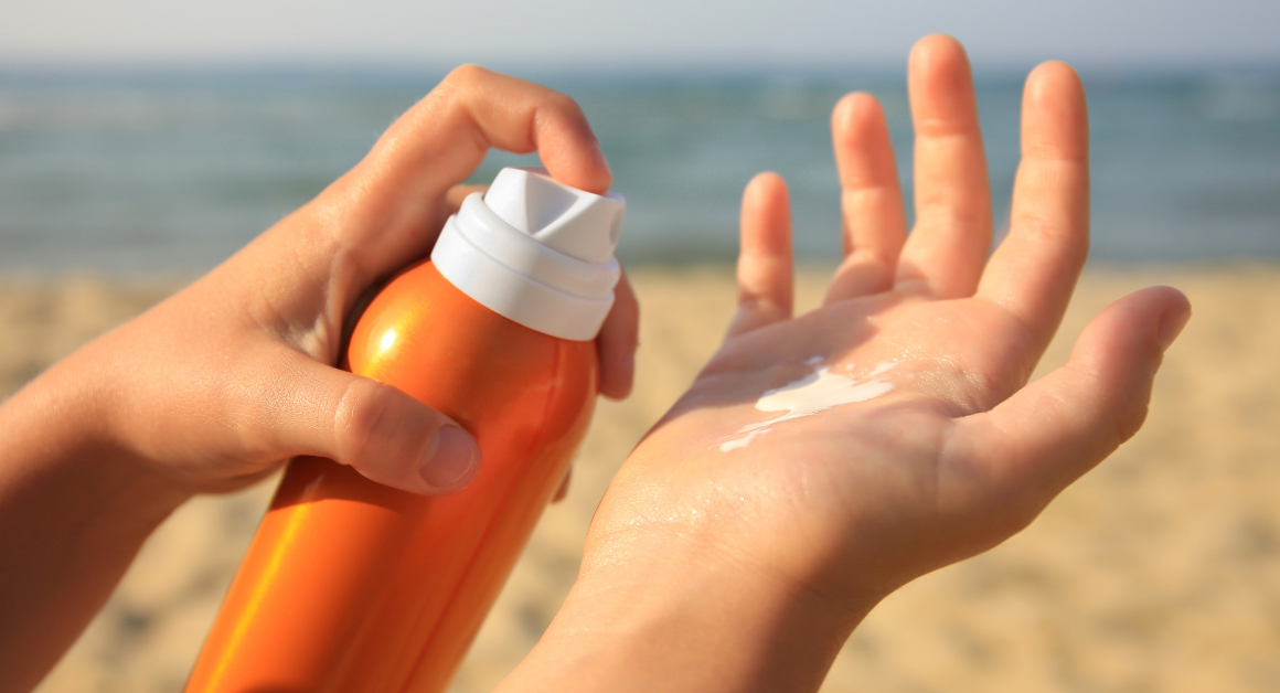 Hands applying sunscreen from an orange bottle on a sandy beach with the ocean in the background, conveying a sense of summer and sun protection.