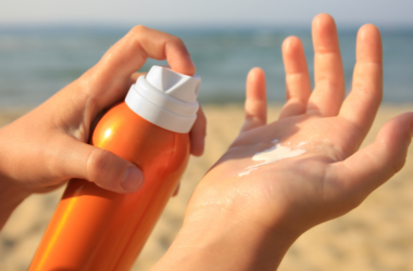 Hands applying sunscreen from an orange bottle on a sandy beach with the ocean in the background, conveying a sense of summer and sun protection.