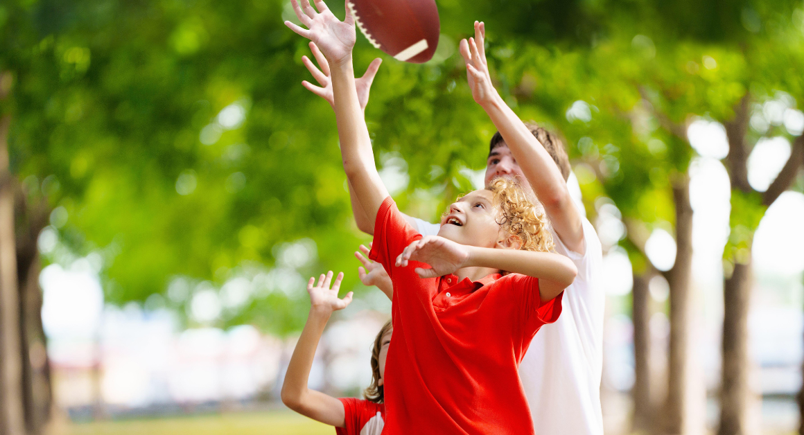 A joyful child reaches to catch a football, surrounded by trees in a park. The scene conveys excitement and playfulness on a sunny day.
