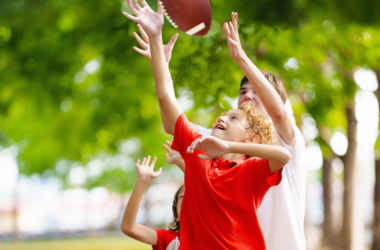 A joyful child reaches to catch a football, surrounded by trees in a park. The scene conveys excitement and playfulness on a sunny day.