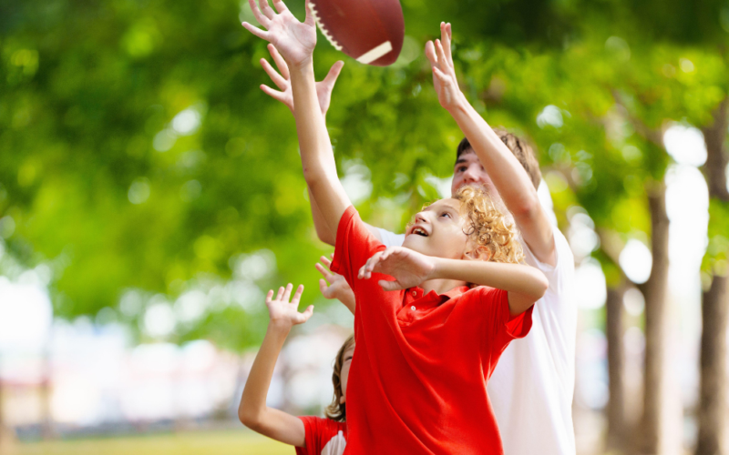 A joyful child reaches to catch a football, surrounded by trees in a park. The scene conveys excitement and playfulness on a sunny day.