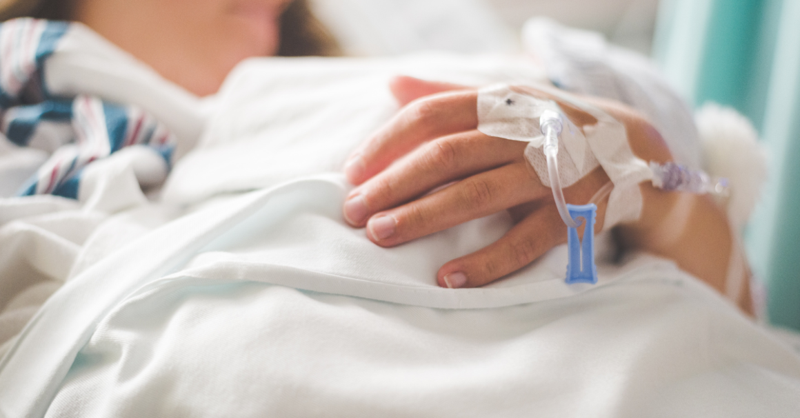 A person in a hospital bed rests their hand on a white blanket, an IV line taped to their hand, representing Chronic Disease Awareness Day.