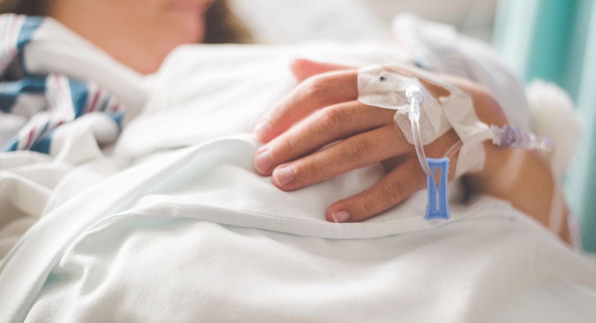A person in a hospital bed rests their hand on a white blanket, an IV line taped to their hand, representing Chronic Disease Awareness Day.