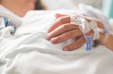 A person in a hospital bed rests their hand on a white blanket, an IV line taped to their hand, representing Chronic Disease Awareness Day.