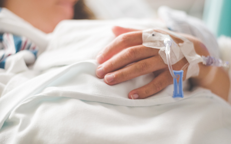 A person in a hospital bed rests their hand on a white blanket, an IV line taped to their hand, representing Chronic Disease Awareness Day.