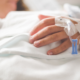 A person in a hospital bed rests their hand on a white blanket, an IV line taped to their hand, representing Chronic Disease Awareness Day.
