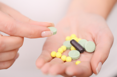 A hand holds various pills: green tablets, yellow round pills, and a black capsule. Another hand picks up a green tablet, conveying a clinical and focused tone.