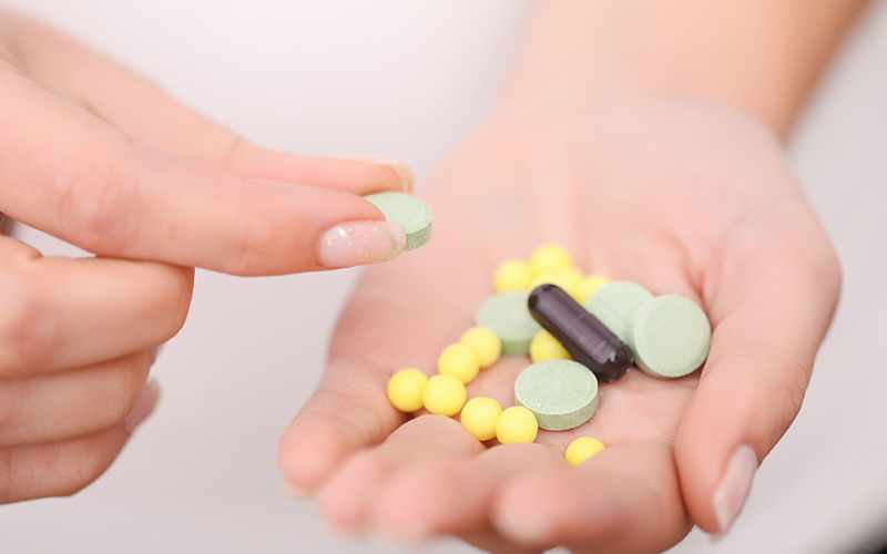 A hand holds various pills: green tablets, yellow round pills, and a black capsule. Another hand picks up a green tablet, conveying a clinical and focused tone.