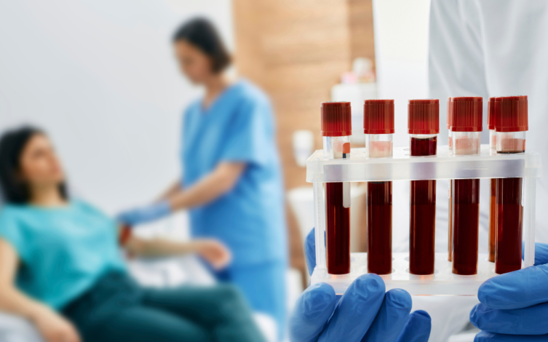 A healthcare professional holding a tray of blood samples in the foreground. In the background, a nurse prepares a patient for a blood draw, creating a clinical and focused atmosphere.