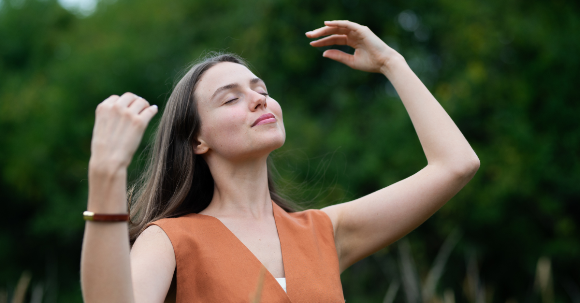 A woman stands outdoors with closed eyes, smiling and raising her arms, enjoying a peaceful moment, representing self-care.
