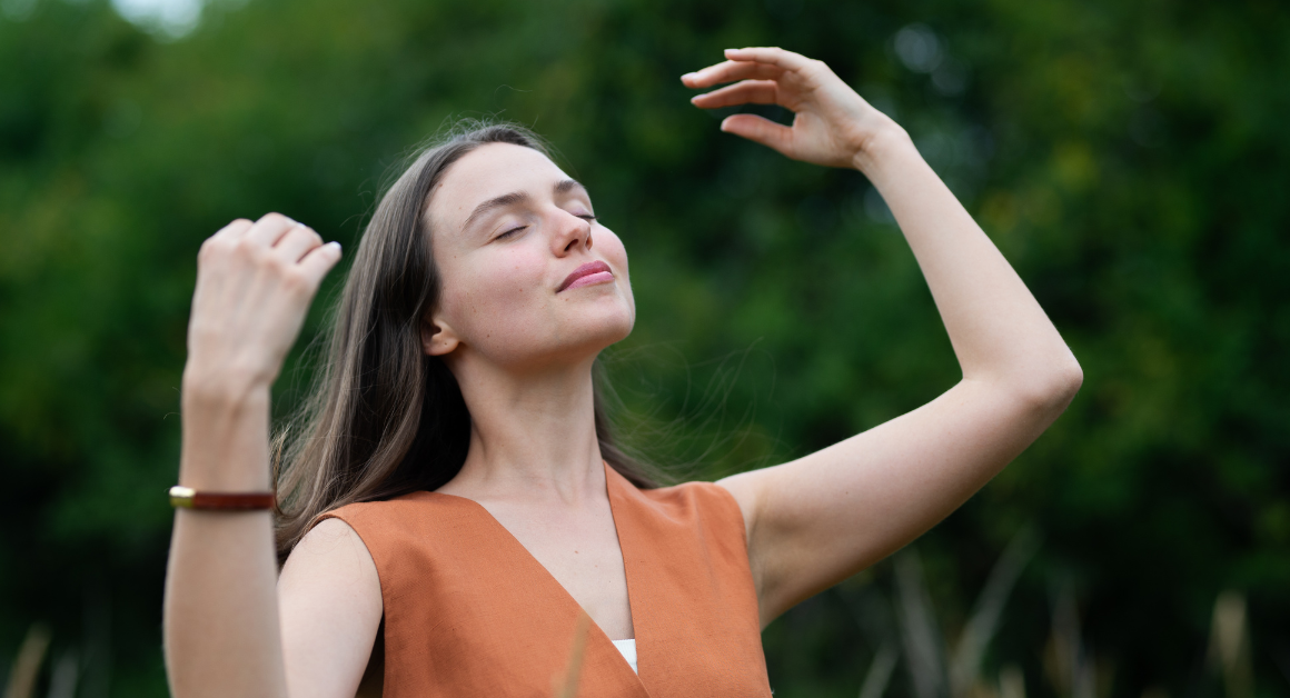 A woman stands outdoors with closed eyes, smiling and raising her arms, enjoying a peaceful moment, representing self-care.