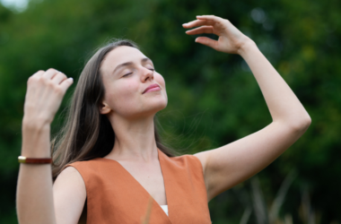 A woman stands outdoors with closed eyes, smiling and raising her arms, enjoying a peaceful moment, representing self-care.
