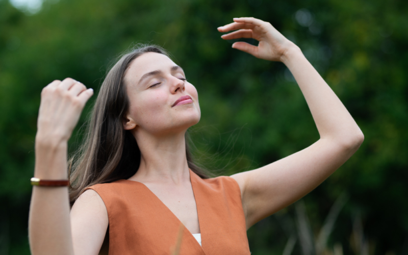 A woman stands outdoors with closed eyes, smiling and raising her arms, enjoying a peaceful moment, representing self-care.