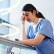 A woman in blue scrubs leans on a railing, looking exhausted and rubbing her forehead, representing a burnout.