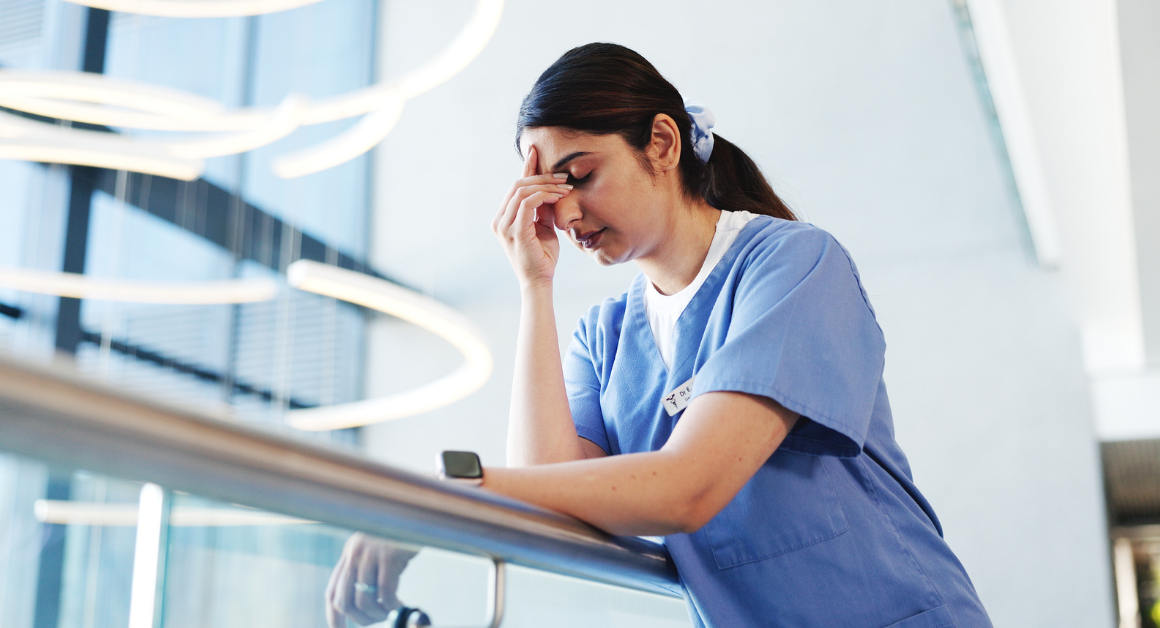 A woman in blue scrubs leans on a railing, looking exhausted and rubbing her forehead, representing a burnout.