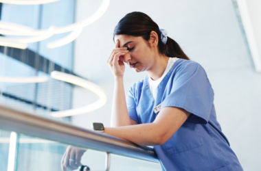 A woman in blue scrubs leans on a railing, looking exhausted and rubbing her forehead, representing a burnout.