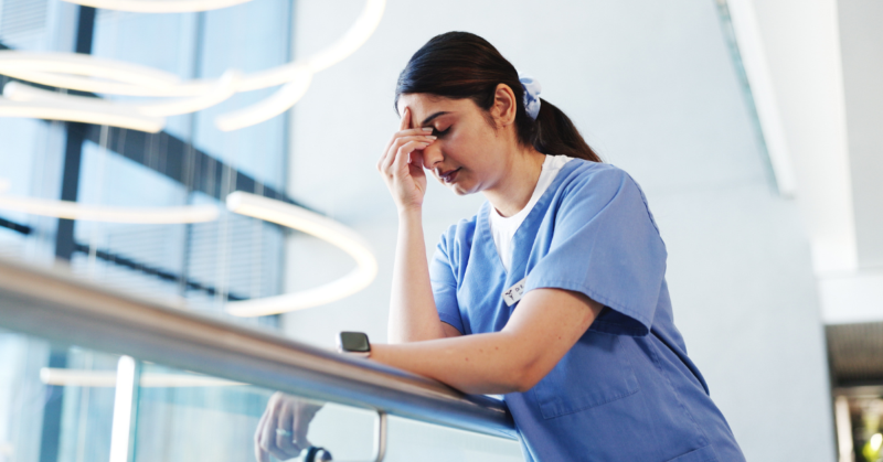 A woman in blue scrubs leans on a railing, looking exhausted and rubbing her forehead, representing a burnout.