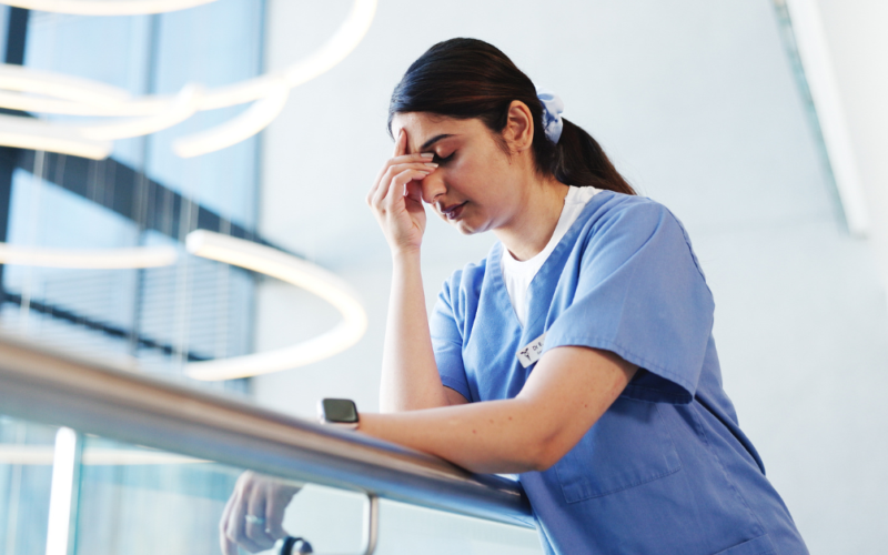 A woman in blue scrubs leans on a railing, looking exhausted and rubbing her forehead, representing a burnout.