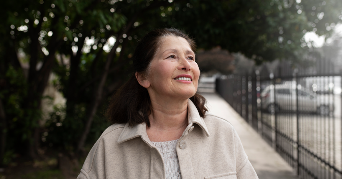 Smiling older woman stands outdoors on a tree-lined sidewalk, looking upward in natural daylight.