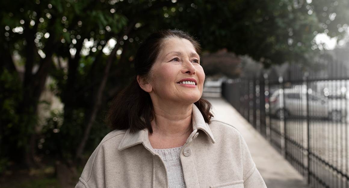 Smiling older woman stands outdoors on a tree-lined sidewalk, looking upward in natural daylight.