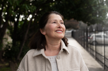 Smiling older woman stands outdoors on a tree-lined sidewalk, looking upward in natural daylight.