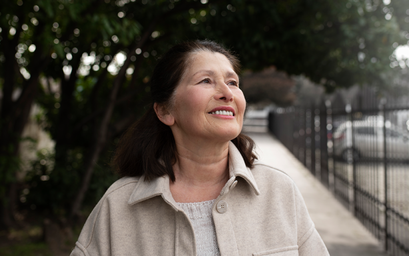 Smiling older woman stands outdoors on a tree-lined sidewalk, looking upward in natural daylight.