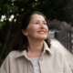 Smiling older woman stands outdoors on a tree-lined sidewalk, looking upward in natural daylight.