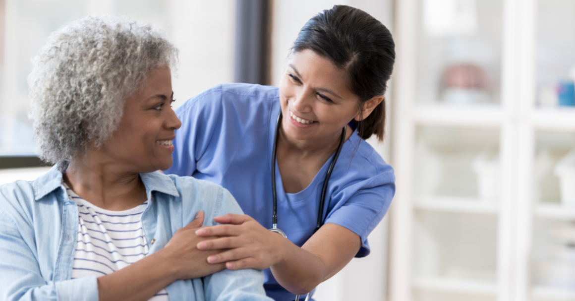 Nurse in blue scrubs smiling and offering reassurance to an older patient during a healthcare visit.