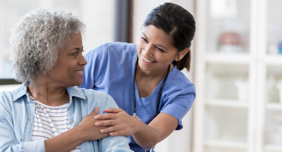 Nurse in blue scrubs smiling and offering reassurance to an older patient during a healthcare visit.