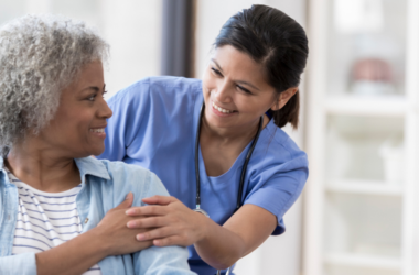 Nurse in blue scrubs smiling and offering reassurance to an older patient during a healthcare visit.