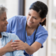 Nurse in blue scrubs smiling and offering reassurance to an older patient during a healthcare visit.