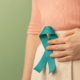 Person holding a teal awareness ribbon against their abdomen, representing ovarian cancer awareness and support.