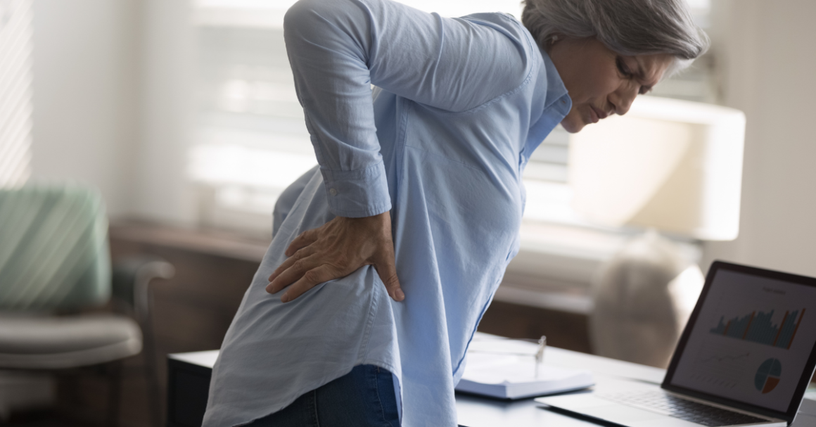An older woman holds her lower back in discomfort while standing beside a desk.