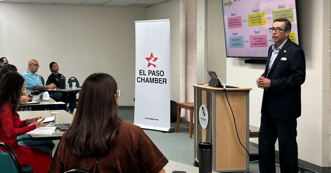 A presenter speaks to attendees during an informational session at the El Paso Chamber, standing beside a screen displaying a step-by-step process while participants listen and take notes.
