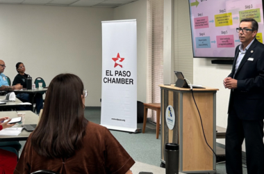 A presenter speaks to attendees during an informational session at the El Paso Chamber, standing beside a screen displaying a step-by-step process while participants listen and take notes.