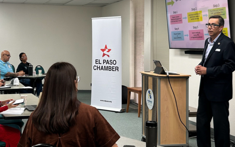 A presenter speaks to attendees during an informational session at the El Paso Chamber, standing beside a screen displaying a step-by-step process while participants listen and take notes.